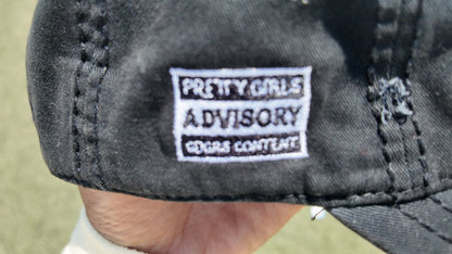 Close-up of a black baseball cap held by a hand, showing a white embroidered patch that reads 'PRETTY GIRLS ADVISORY GDGRS CONTENT.